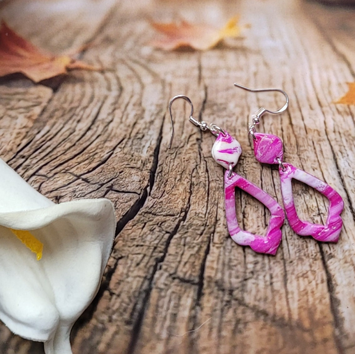 Vue d’ensemble des boucles d’oreilles Rétro-chic à pendentifs allongés ajourés, disposées près d’un arum blanc sur un décor boisé.
