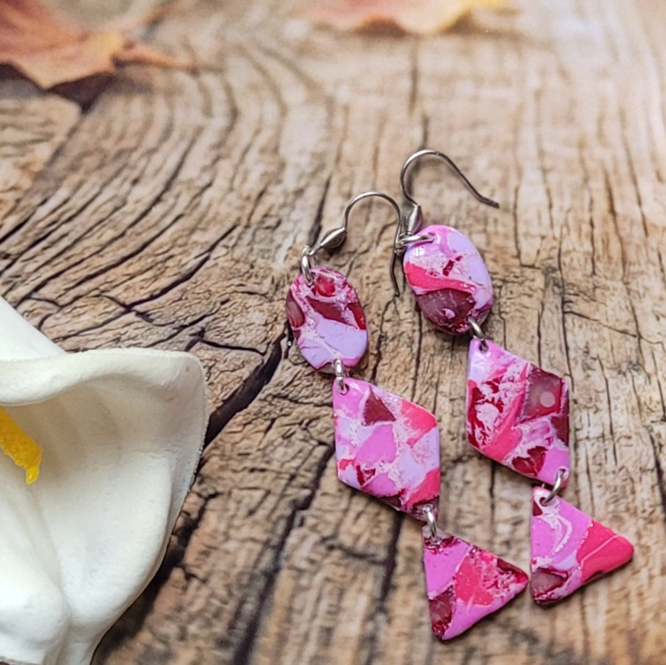 Boucles d’oreilles Rétro-chic composées de trois formes articulées, ovale, losange et triangle, aux motifs roses et bordeaux près d’un arum blanc