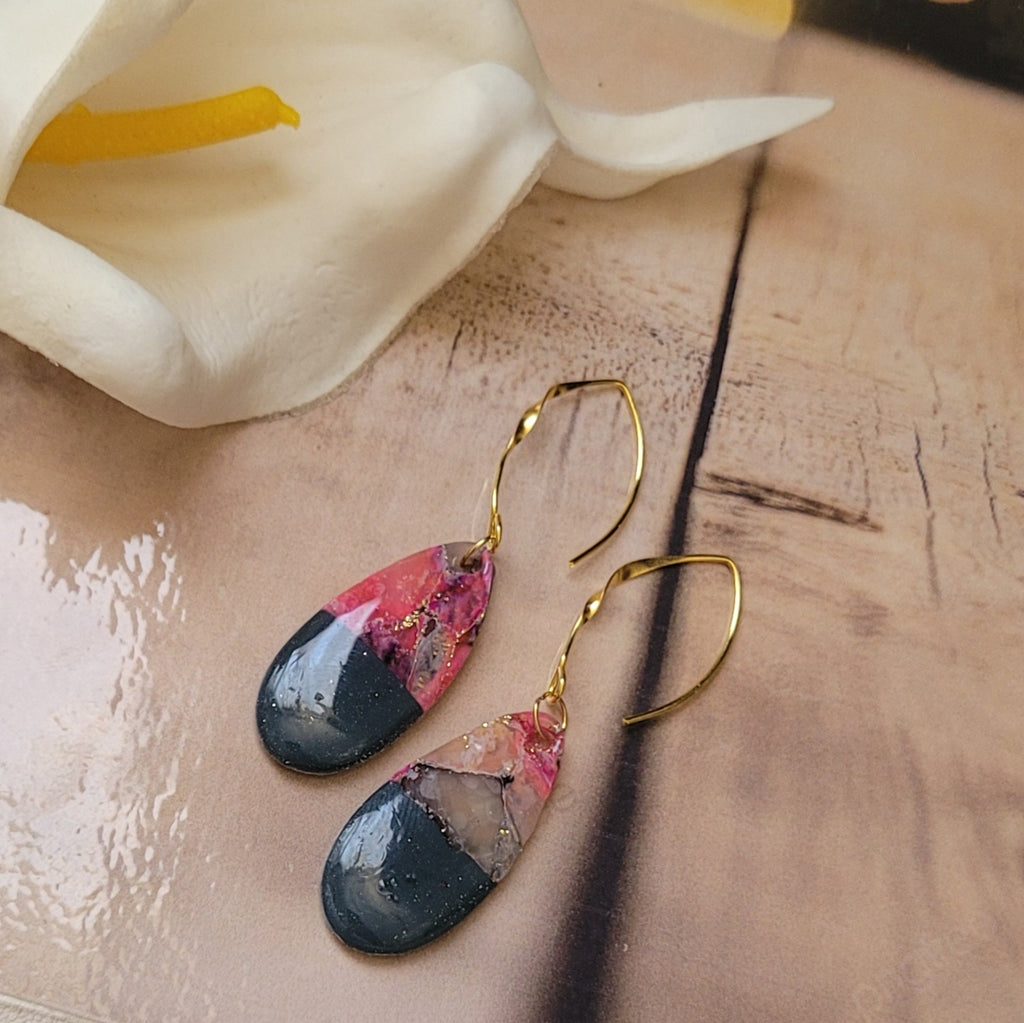 Paire de boucles d’oreilles Éclipse aux pendentifs en goutte roses, translucides et gris foncé, avec de longues attaches dorées, sur un décor de bois clair près d’un arum blanc.