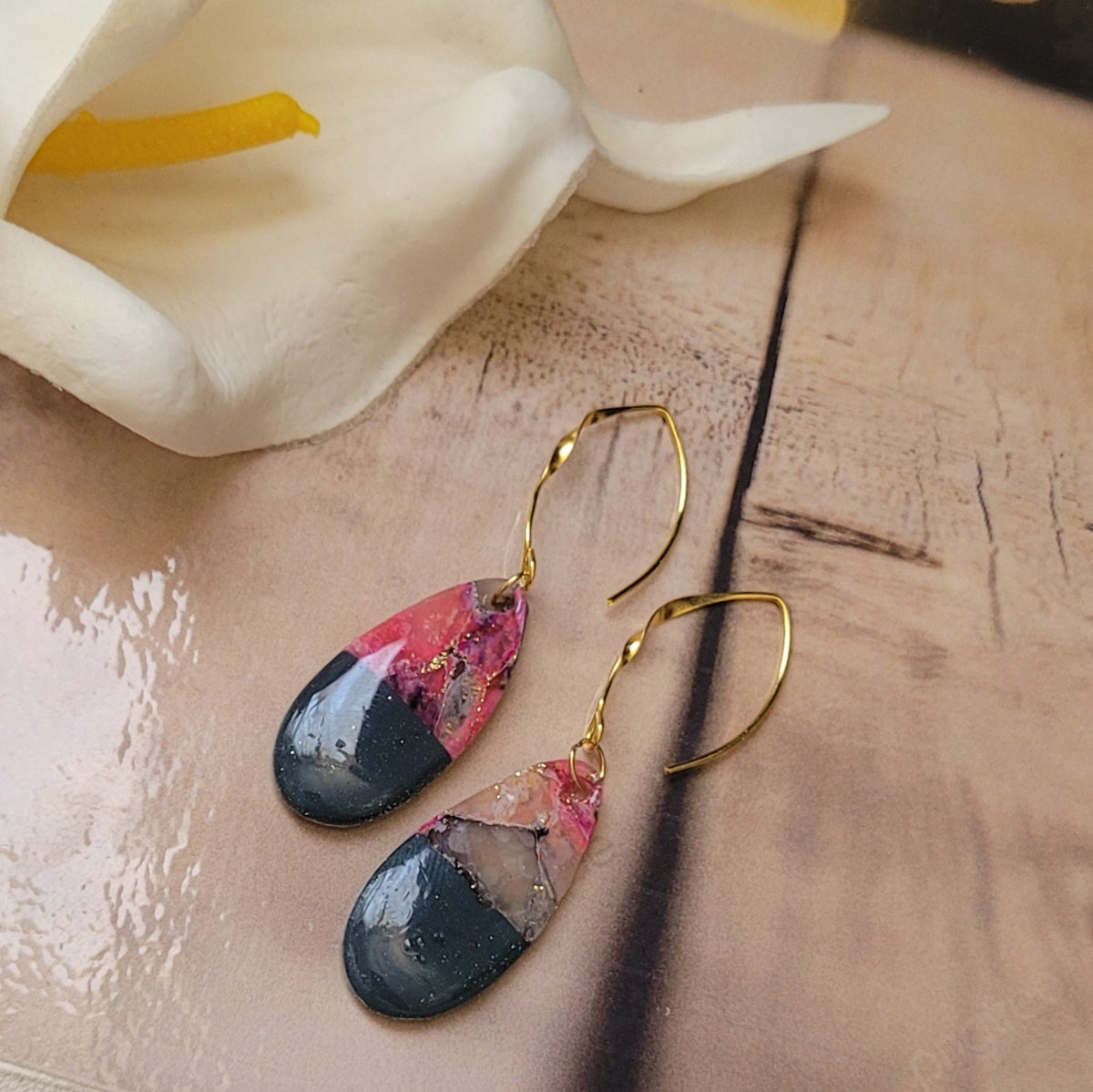 Paire de boucles d’oreilles Éclipse aux pendentifs en goutte roses, translucides et gris foncé, avec de longues attaches dorées, sur un décor de bois clair près d’un arum blanc.