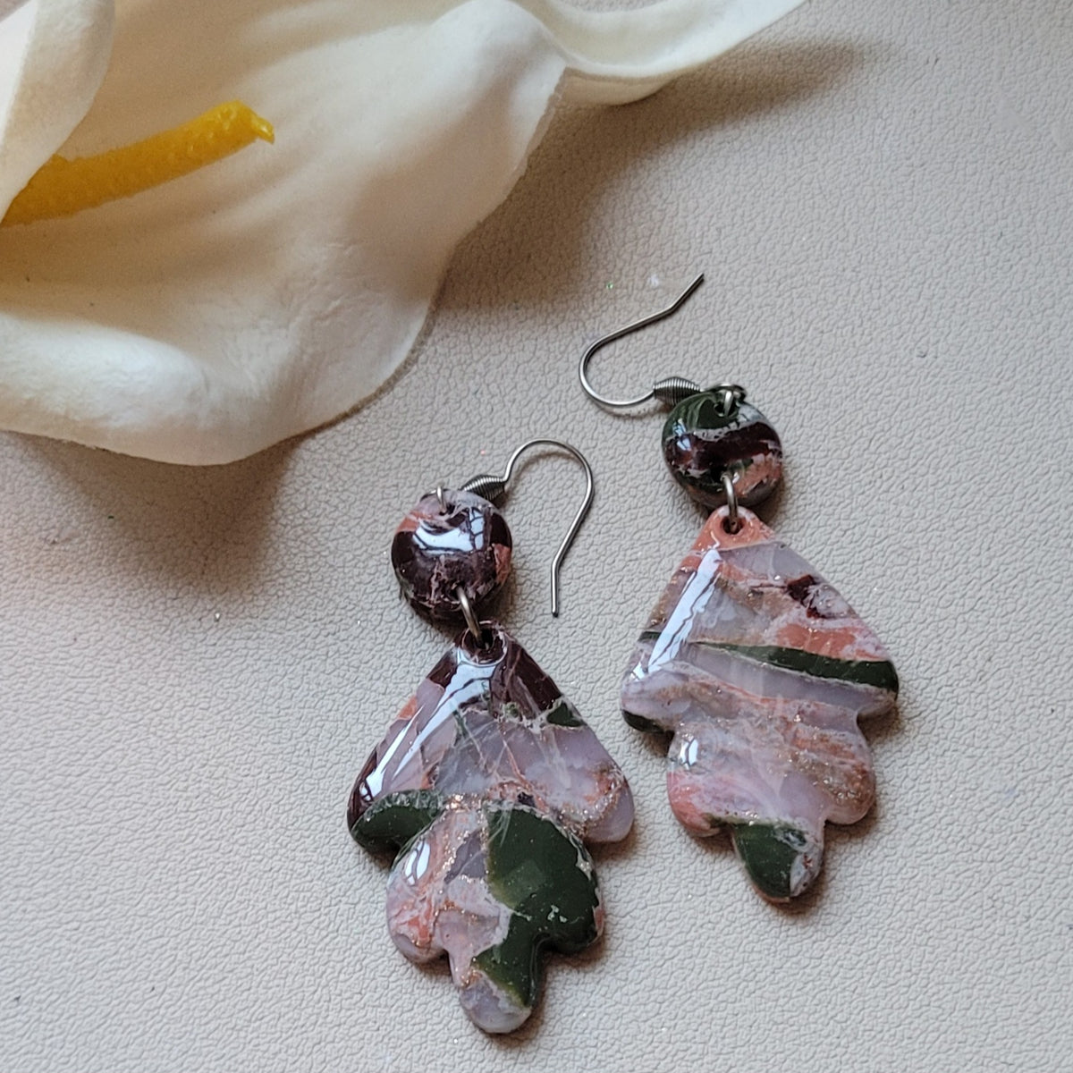 Paire de boucles d’oreilles Flora Feuille aux motifs roses, mauves et vert foncé, avec un élément rond et un pendentif végétal stylisé, près d’un arum blanc sur fond clair texturé.
