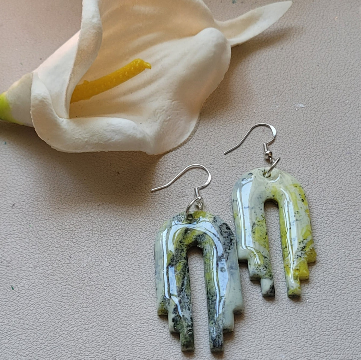  Paire de boucles d’oreilles Empire en forme d’arches ajourées, marbrées de blanc, de jaune-vert et de noir, sur fond clair texturé près d’un arum blanc.