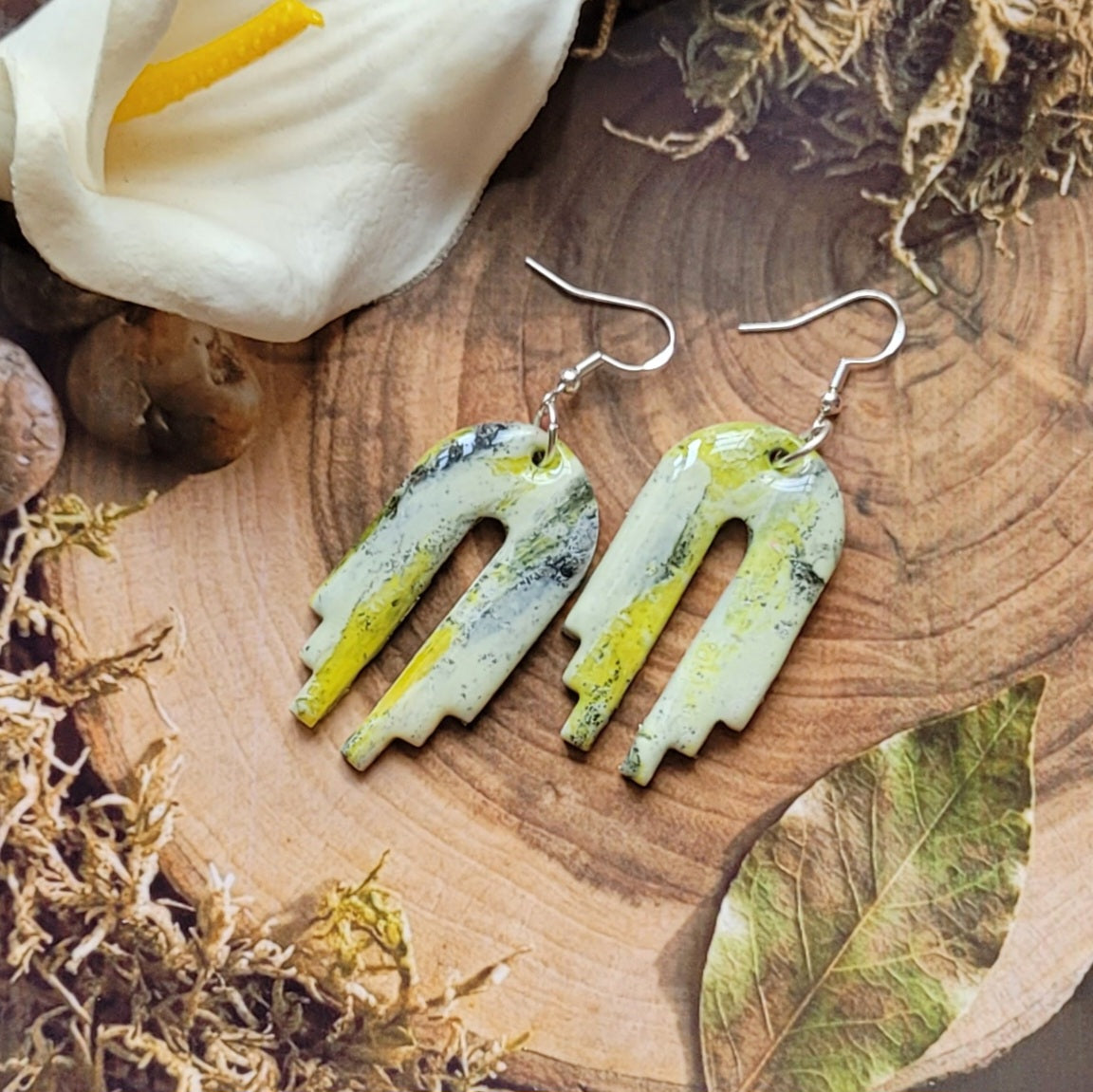  Paire de boucles Empire posée sur une tranche de bois, entourée de végétaux séchés, d’une feuille et d’un arum blanc.