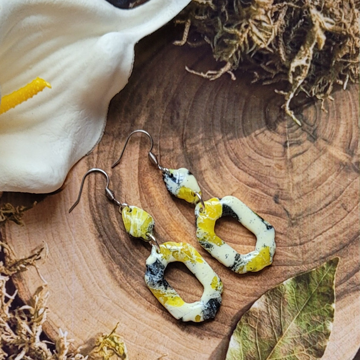 Paire de boucles d’oreilles Aquarelle vanillée aux motifs crème, jaunes et noirs, posée sur une tranche de bois près d’un arum blanc et de végétaux séchés.