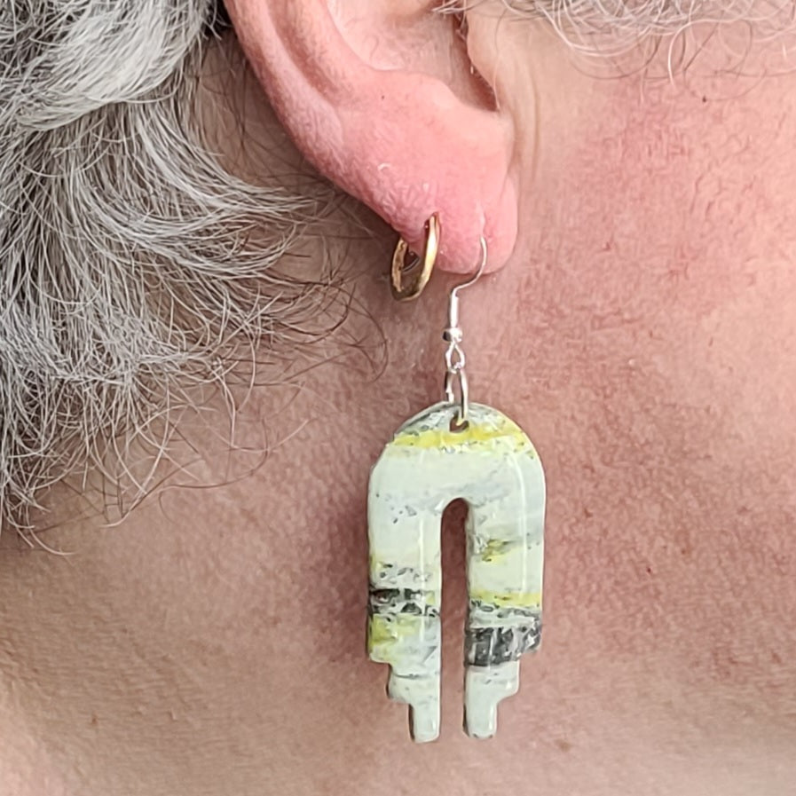  Boucle d’oreille Empire portée, montrant son pendentif en arche ajourée aux nuances claires, jaune-vert et noires et son crochet argenté.