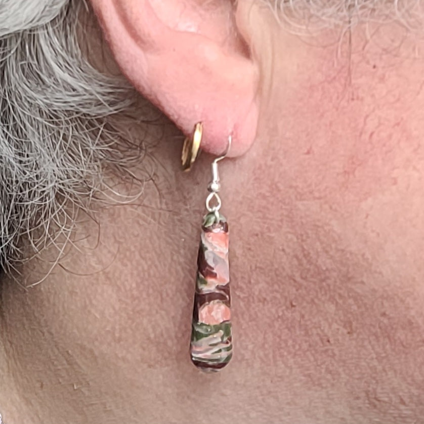 Boucle d’oreille Flora Longues portée, avec un pendentif fin et allongé aux marbrures roses, mauves et vert foncé et un crochet argenté