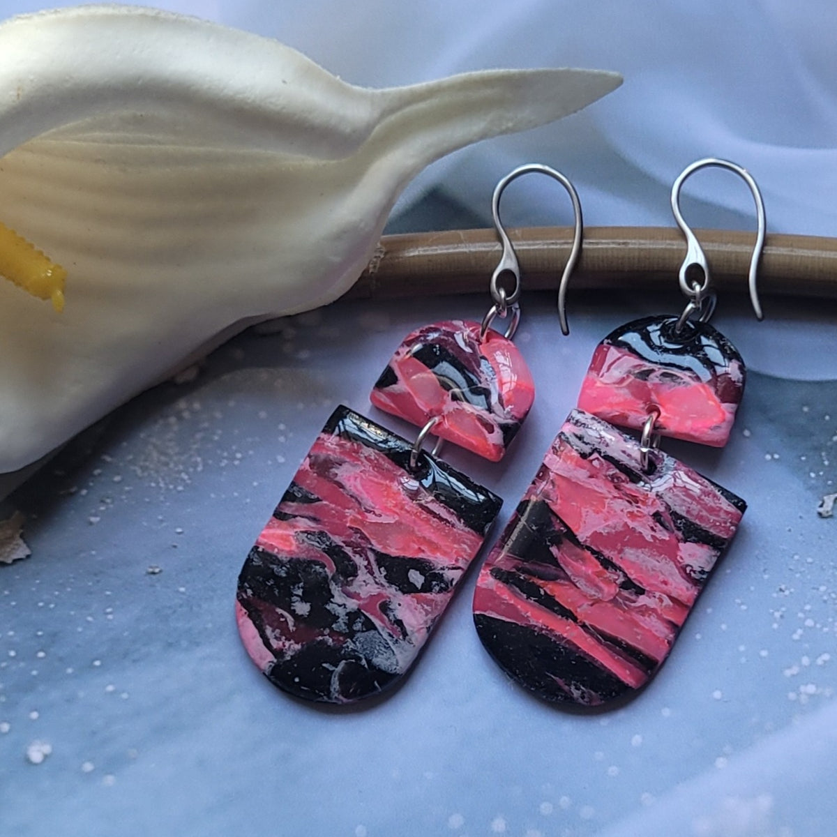  Boucles d’oreilles pendantes Nyméria en pâte polymère et résine, marbrées rose et noir, photographiées à plat.