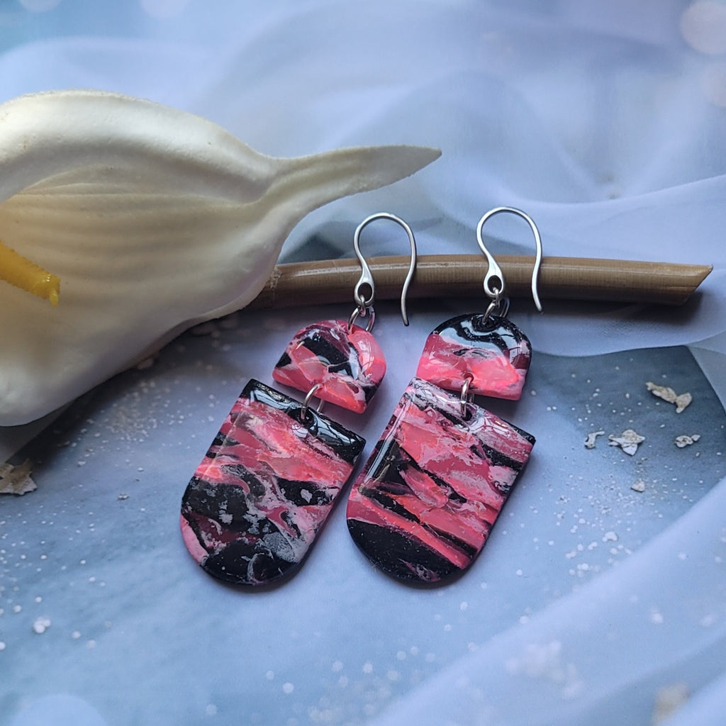  Boucles d’oreilles pendantes Nyméria en pâte polymère et résine, marbrées rose et noir, photographiées à plat.